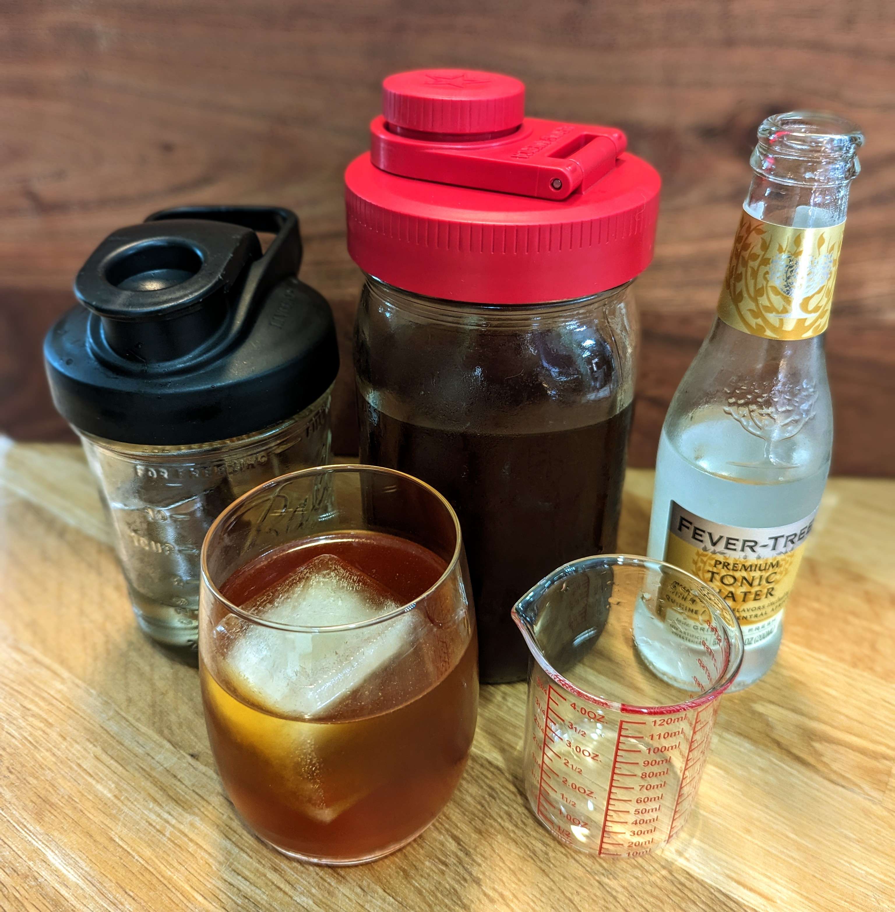Coffee tonic ingredients: a 1-pint mason jar containing simple syrup, a 1-quart mason jar of cold brew coffee, a small bottle of Fever-Tree Tonic, and a glass of prepared coffee tonic with ice.